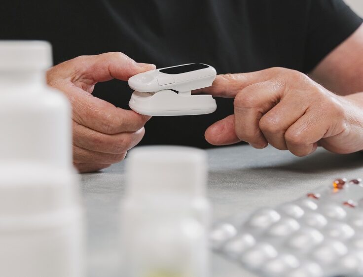 Portrait of one senior unrecognizable woman who measures her pulse on her finger by herself with the help of the device, sitting at the gray table with pills in bottles in the room during the daytime, bottom side view very close-up. Concept of time of using technology, retirement lifestyle.