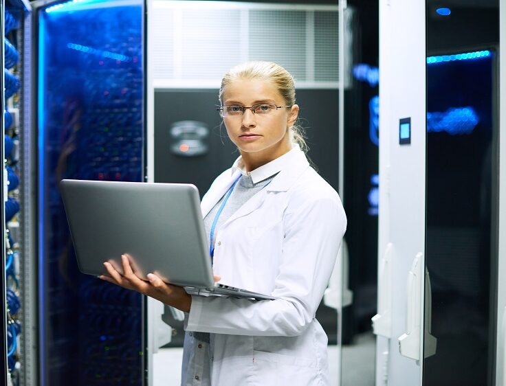 Portrait of young woman wearing lab coat working with supercomputer standing in server room holding laptop